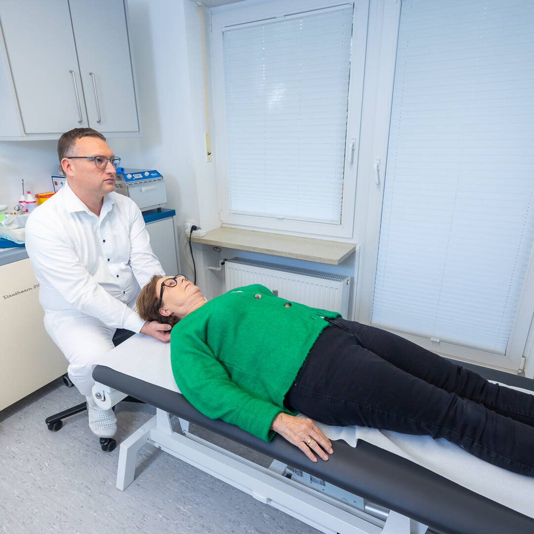 Aufnahme von schräg oben einer älteren, liegenden Frau im grünen Pullover, welche am Halswirbel von Dr.Mattias Hartmann untersucht wird