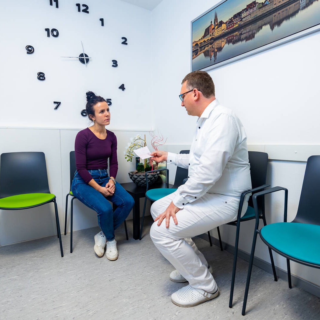 Dr. Hartmann und eine Patientin unterhalten sich sitzend im Wartezimmer der Praxis