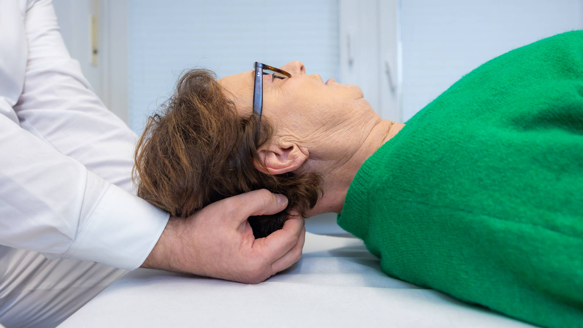 Seitliche Nahaufnahme Kopf und Schultern einer älteren, liegenden Frau im grünen Pullover, welche am Halswirbel untersucht wird