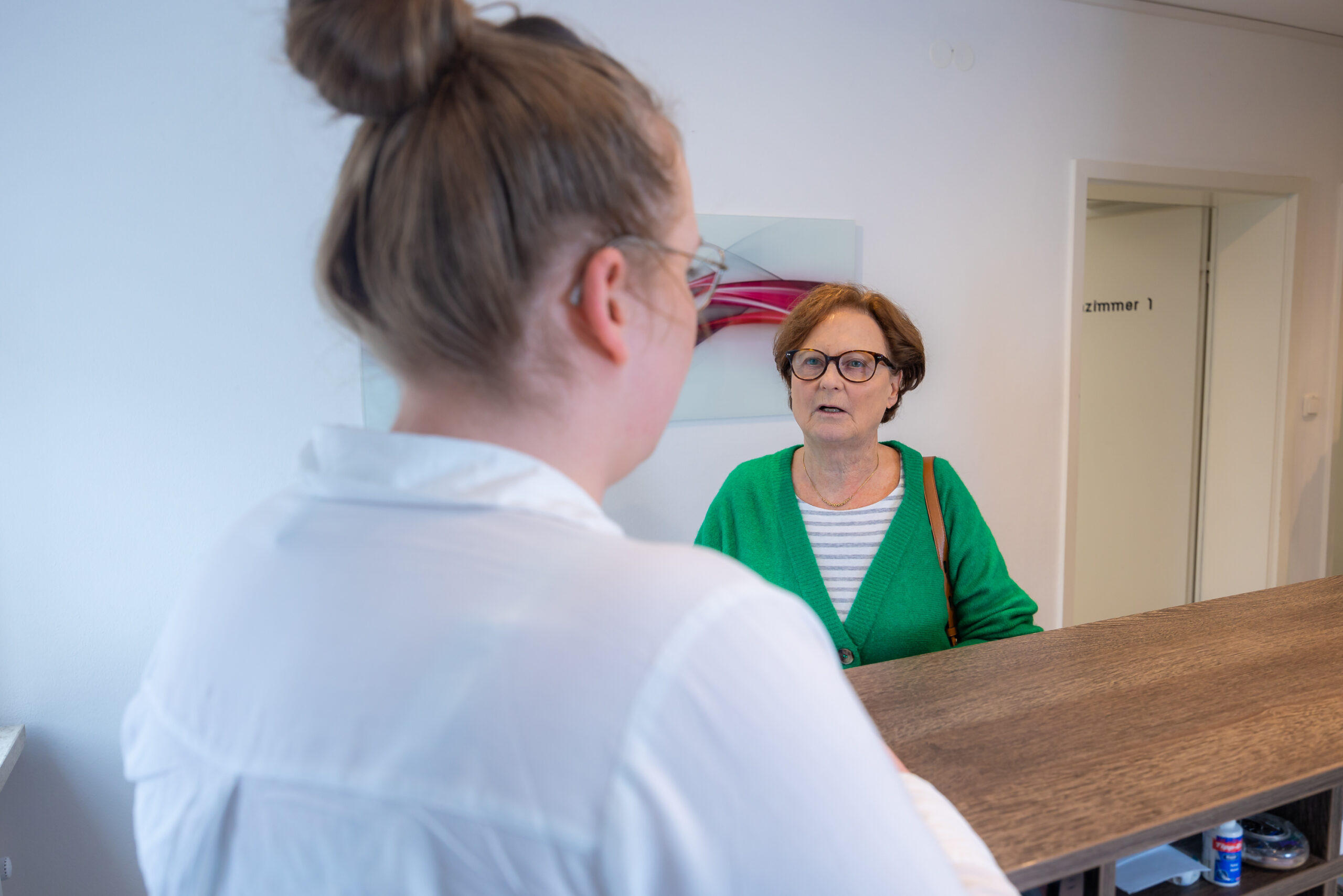 Nahaufnahme von hinten einer Angestellten an der Theke an welcher eine ältere Patientin im grünen Pullover steht