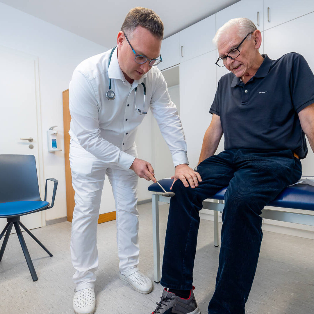 Dr. Matthias Hartmann untersucht einen älteren Man am Knie, welcher auf einer blauen Liege sitzt
