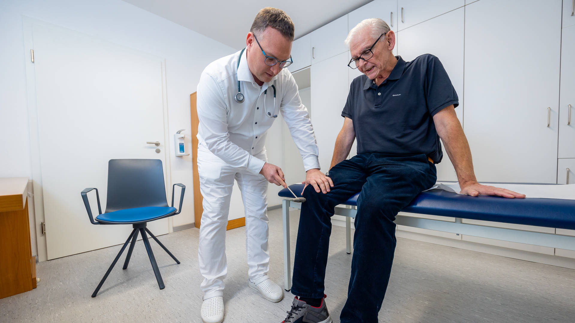 Dr. Matthias Hartmann untersucht einen älteren Man am Knie, welcher auf einer blauen Liege sitzt